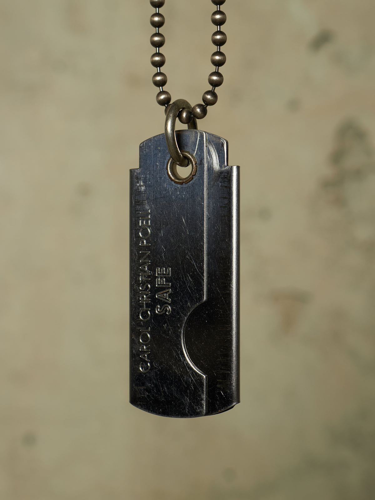 A dark gunmetal dog tag pendant necklace with a ball chain, featuring the engraved text 'GARO CHAD PENIS SAFE' on a textured beige background.