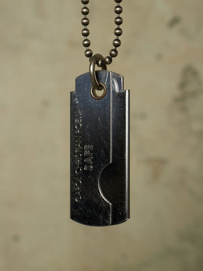 A dark gunmetal dog tag pendant necklace with a ball chain, featuring the engraved text 'GARO CHAD PENIS SAFE' on a textured beige background.