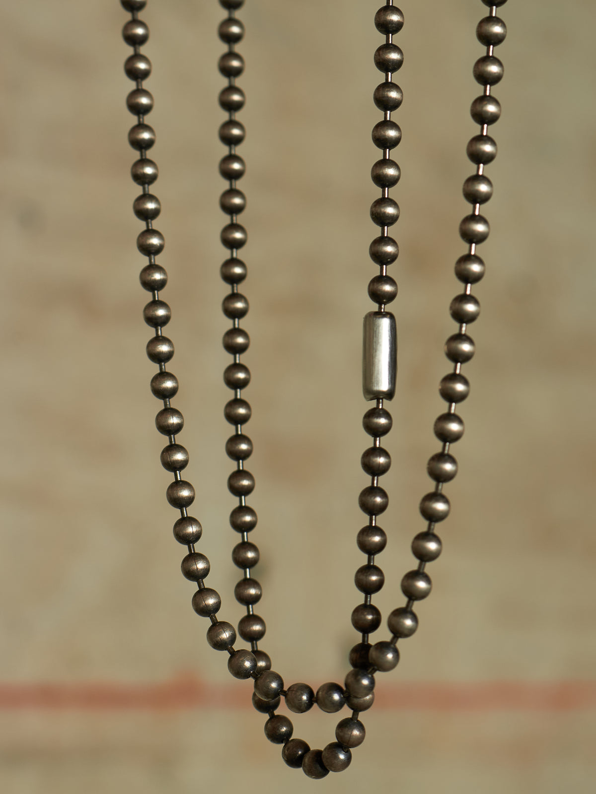 A close-up of a multi-strand beaded necklace with dark metallic bronze beads and a single silver cylindrical accent bead, set against a warm beige background.