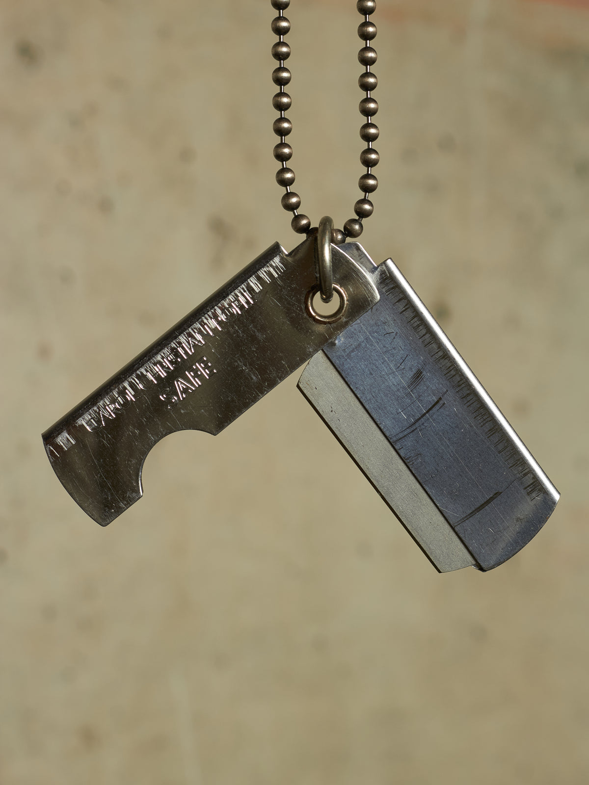 A silver metal dog tag necklace with a razor blade-shaped tag engraved with 'TO THOSE WHO WILL LOOK', hanging from a black ball chain against a textured beige background.
