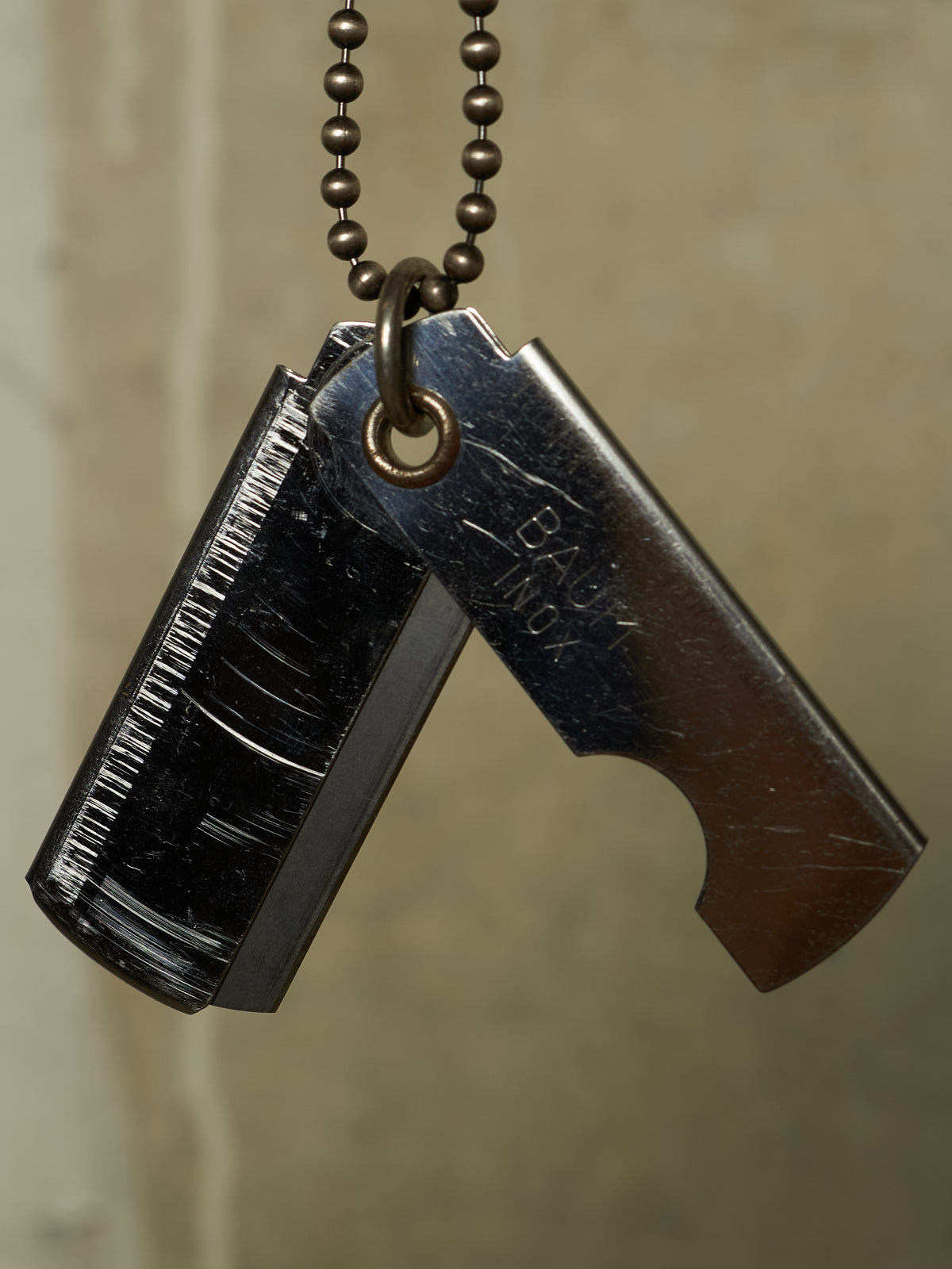 A close-up of a black metal utility knife pendant necklace with a matching ball chain, shown against a neutral beige background.