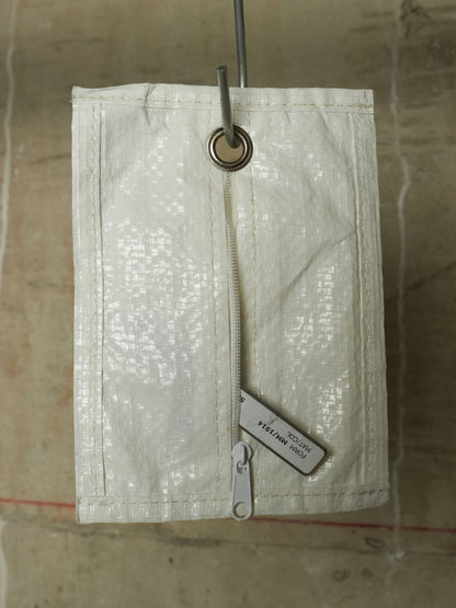 An off-white woven canvas utility pouch with a vertical zipper and a metal grommet at the top, hanging against a textured concrete wall.