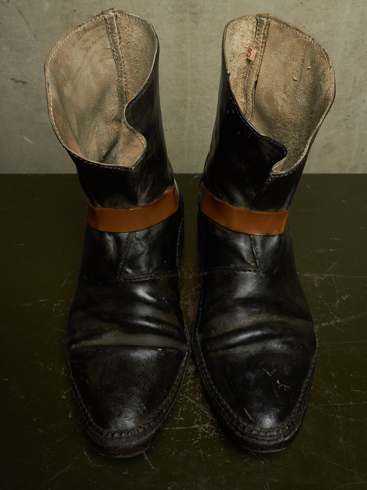 A pair of worn black leather ankle boots with tan leather bands around the upper shaft, photographed on a dark surface against a textured gray wall.