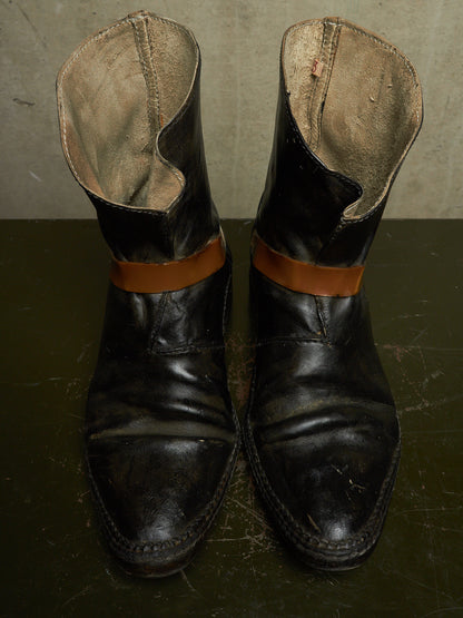 A pair of worn black leather ankle boots with tan leather bands around the upper shaft, photographed on a dark surface against a textured gray wall.