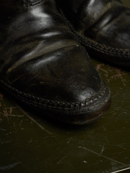 A close-up of a worn black leather boot toe showing the textured leather, braided welt stitching, and scuffed sole, resting on a dark, scratched surface.