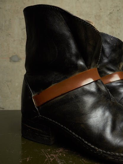 A close-up of a black leather ankle boot with a contrasting brown leather strap detail across the side, resting on a dark surface against a textured concrete wall.