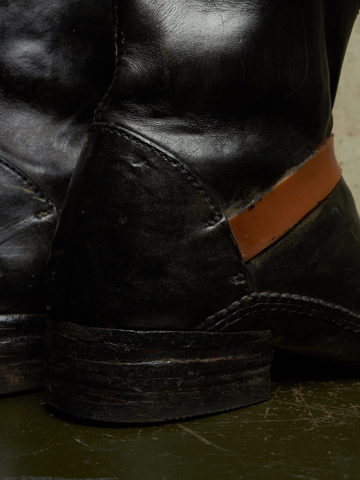 A close-up of a black leather boot with a prominent tan leather heel tab, resting on a dark green surface.