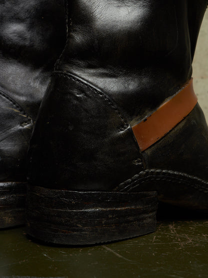A close-up of a black leather boot with a prominent tan leather heel tab, resting on a dark green surface.