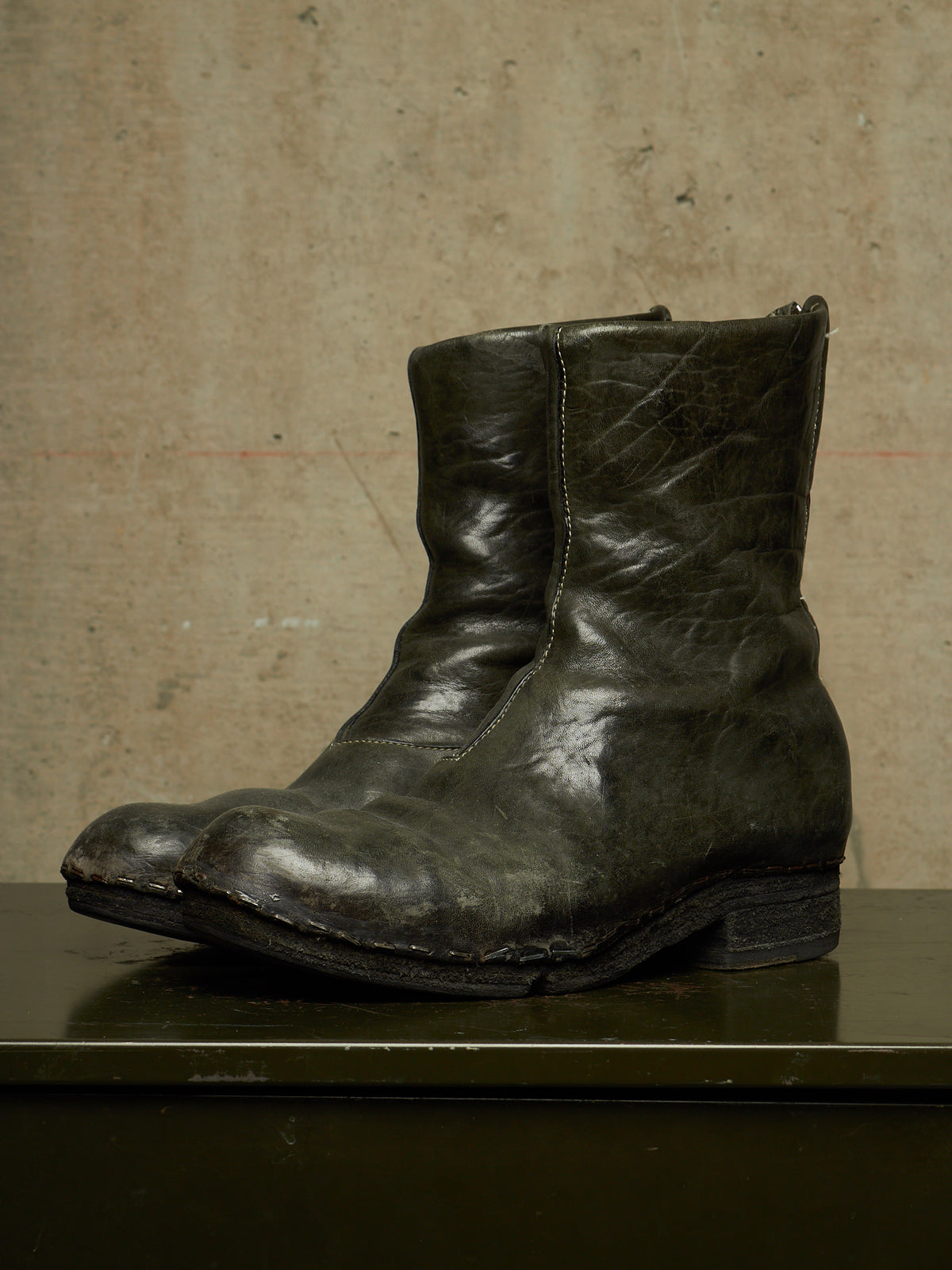 A single dark distressed leather ankle boot with a rugged, worn finish, displayed on a dark surface against a textured concrete wall.