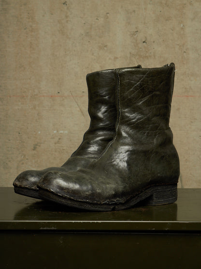 A single dark distressed leather ankle boot with a rugged, worn finish, displayed on a dark surface against a textured concrete wall.