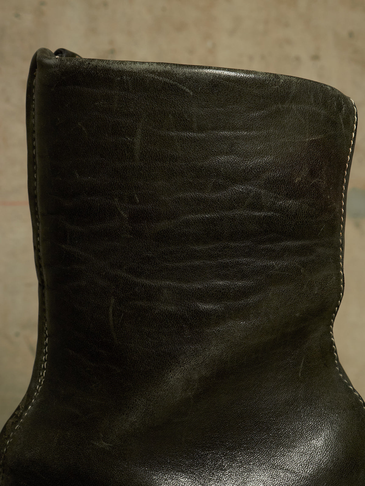 A close-up of a dark olive green leather boot showing the textured leather surface, stitching, and a portion of the sole.
