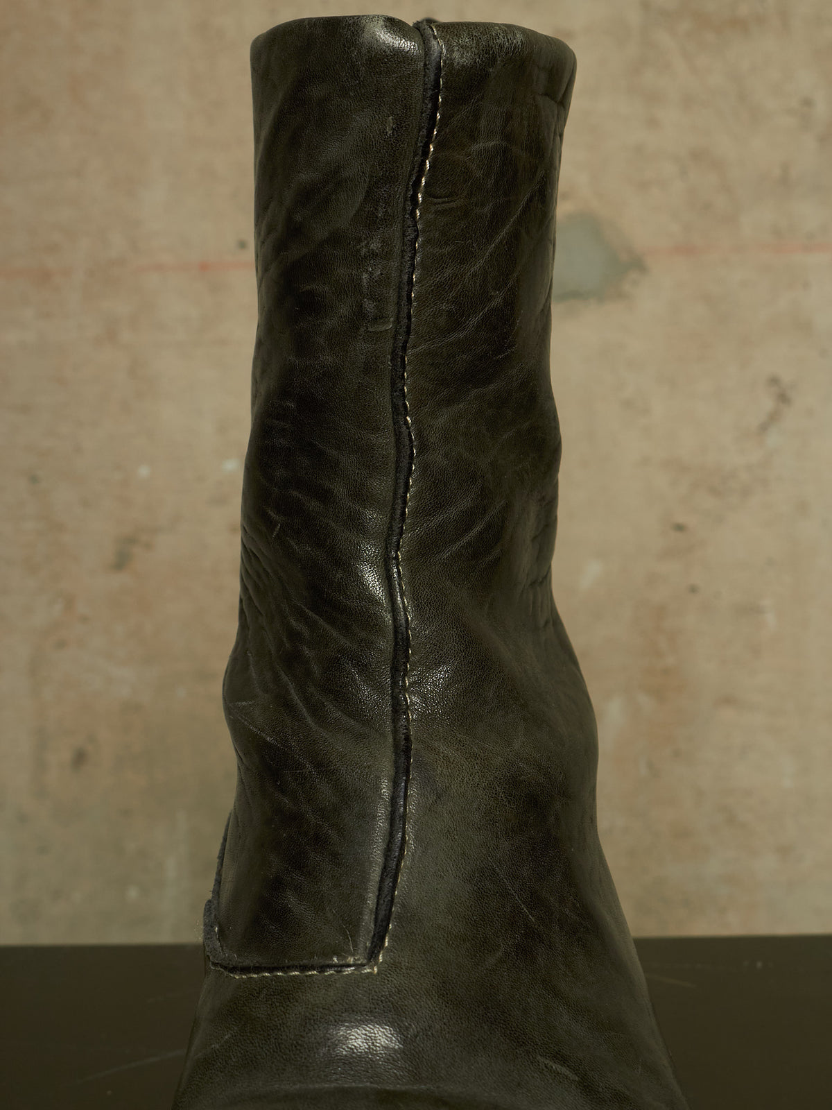 A close-up of a dark distressed leather ankle boot, showing the side stitching and textured leather surface against a neutral concrete background.