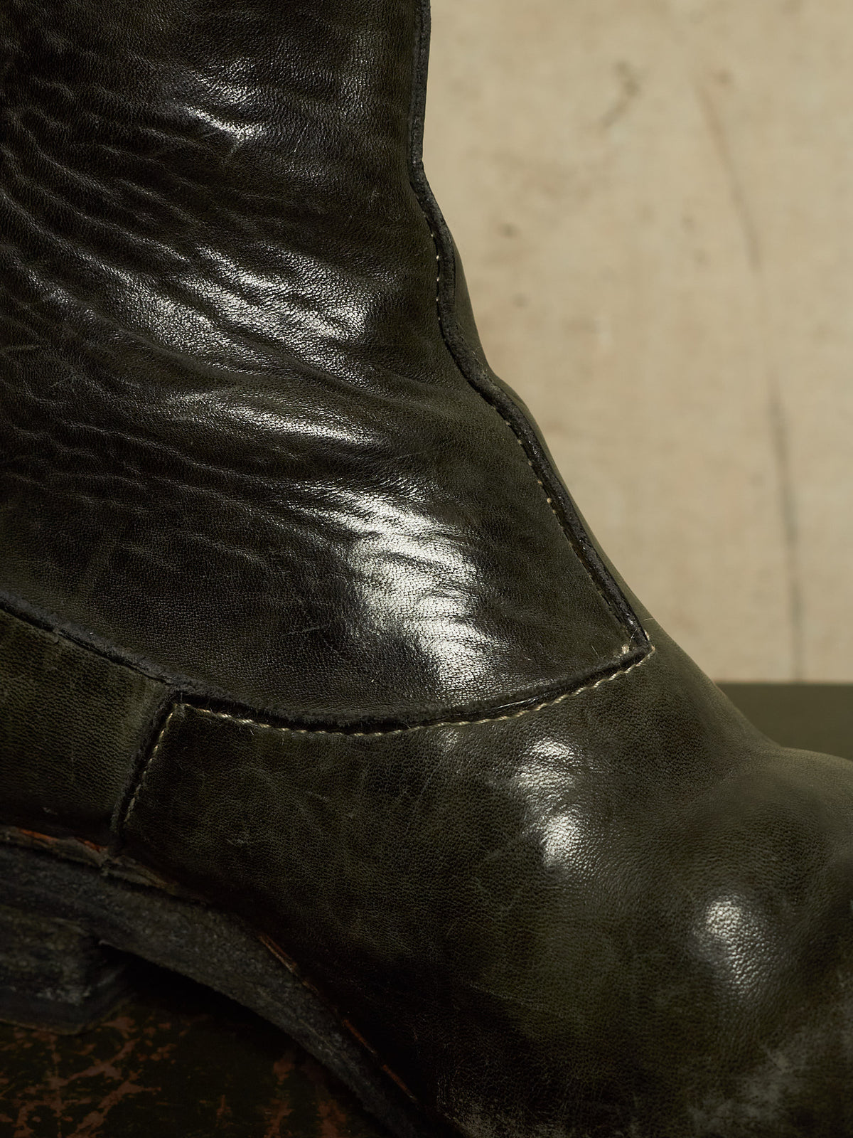 Close-up of a dark distressed leather boot showing the textured leather grain, stitching, and a low heel against a neutral beige background.