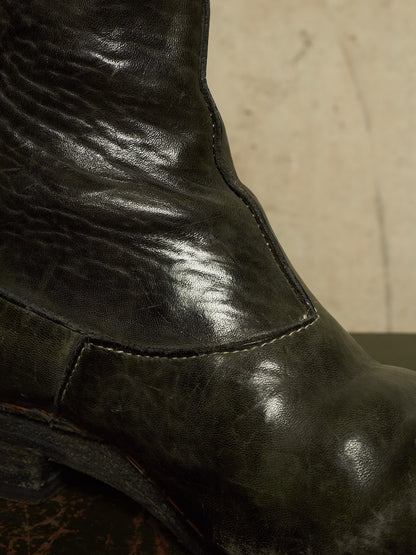 Close-up of a dark distressed leather boot showing the textured leather grain, stitching, and a low heel against a neutral beige background.