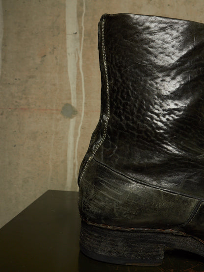 A close-up of a worn black leather boot with prominent creasing and scuffing, positioned on a dark surface against a textured beige wall.