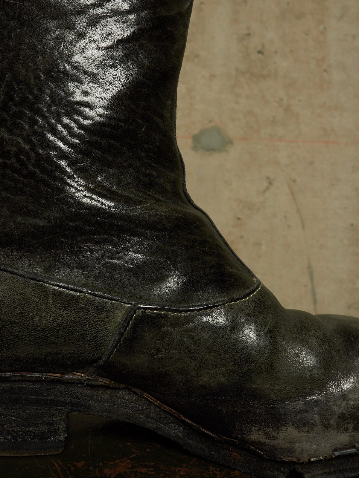 A close-up of a worn black leather boot with a dark sole, set against a textured beige concrete background.