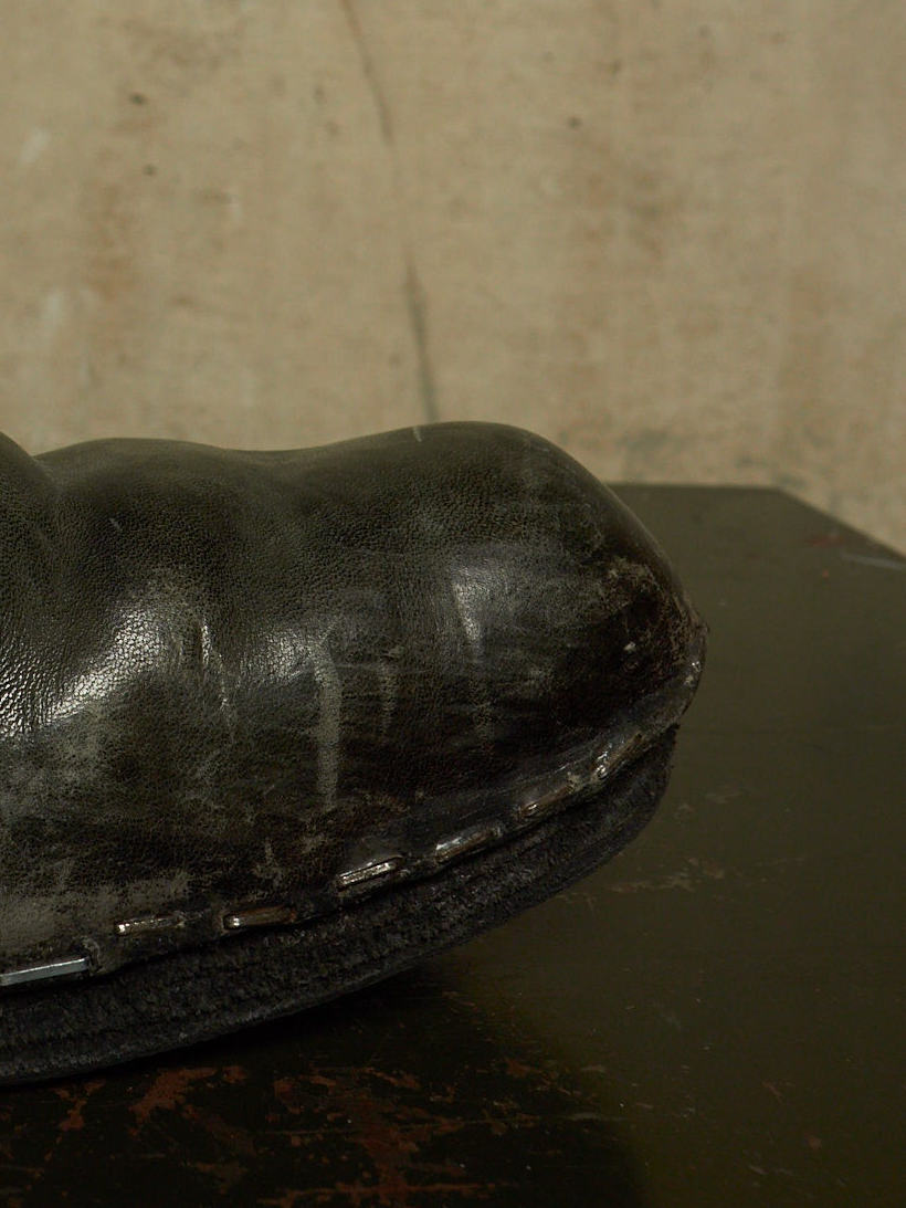 A close-up of a worn black leather boot or shoe resting on a dark surface against a textured beige wall.