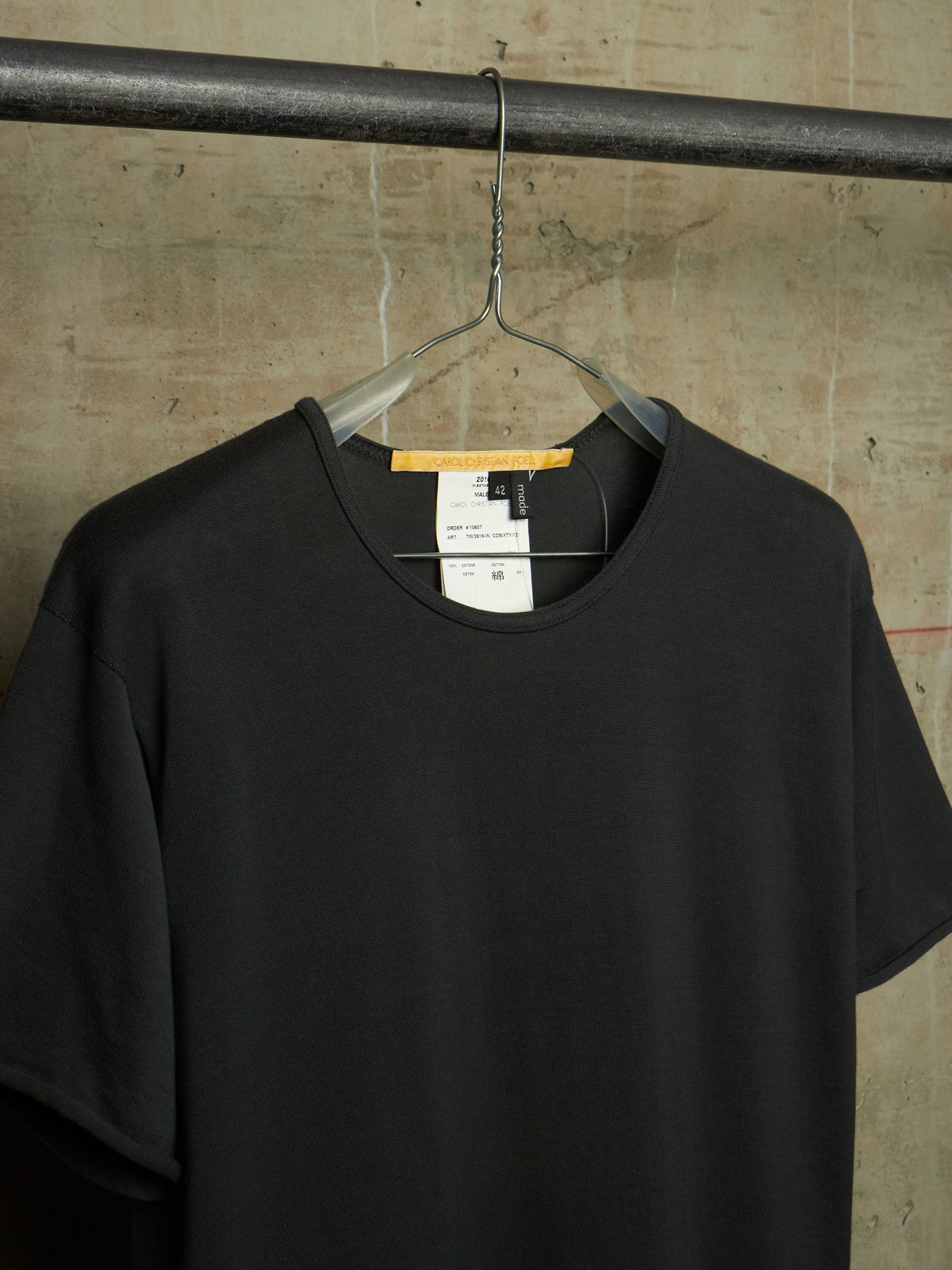 A black short-sleeve crewneck t-shirt hanging on a clear plastic hanger from a dark metal rod, set against a textured concrete wall.