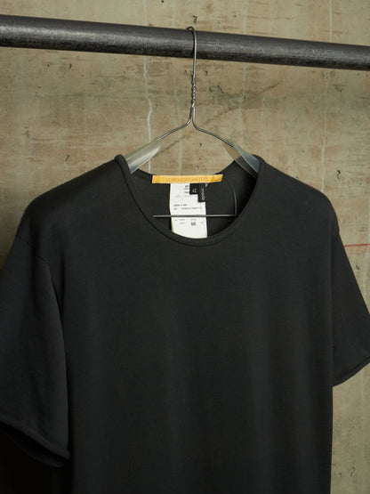 A black short-sleeve crewneck t-shirt hanging on a clear plastic hanger from a dark metal rod, set against a textured concrete wall.