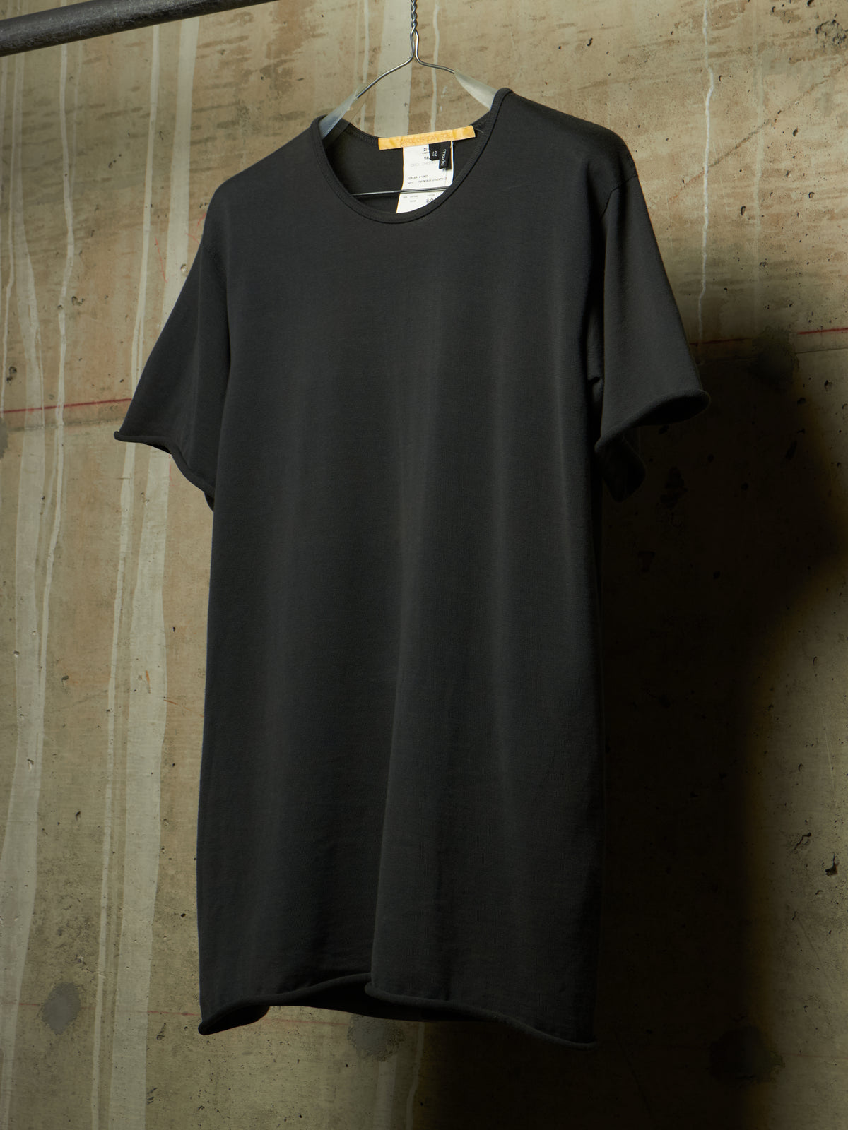 A black short-sleeve crewneck t-shirt hanging on a metal hanger against a textured concrete wall.