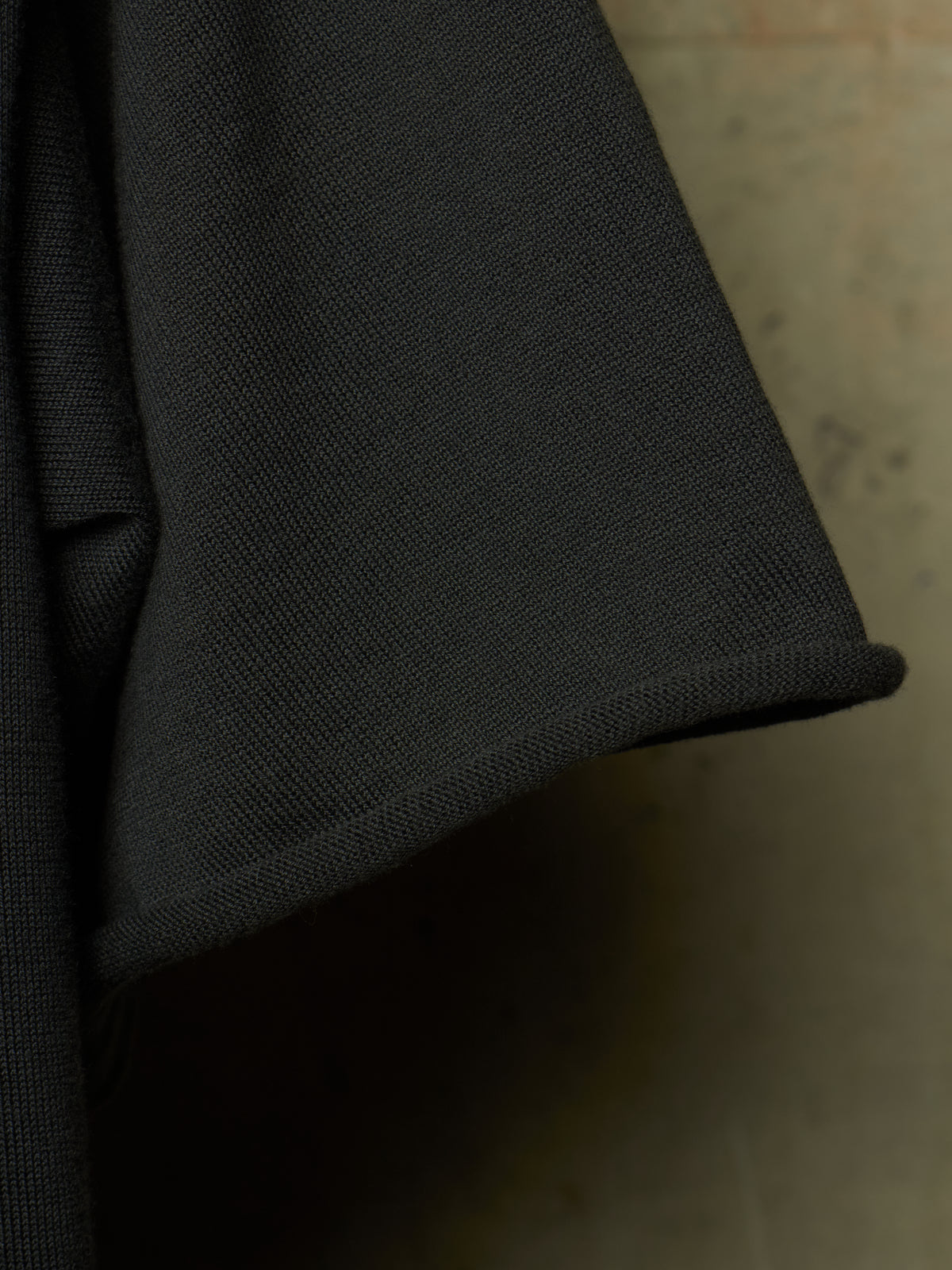 Close-up detail of a black textured fabric garment, showing the weave and a rolled cuff edge against a neutral concrete-like background.