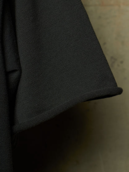 Close-up detail of a black textured fabric garment, showing the weave and a rolled cuff edge against a neutral concrete-like background.