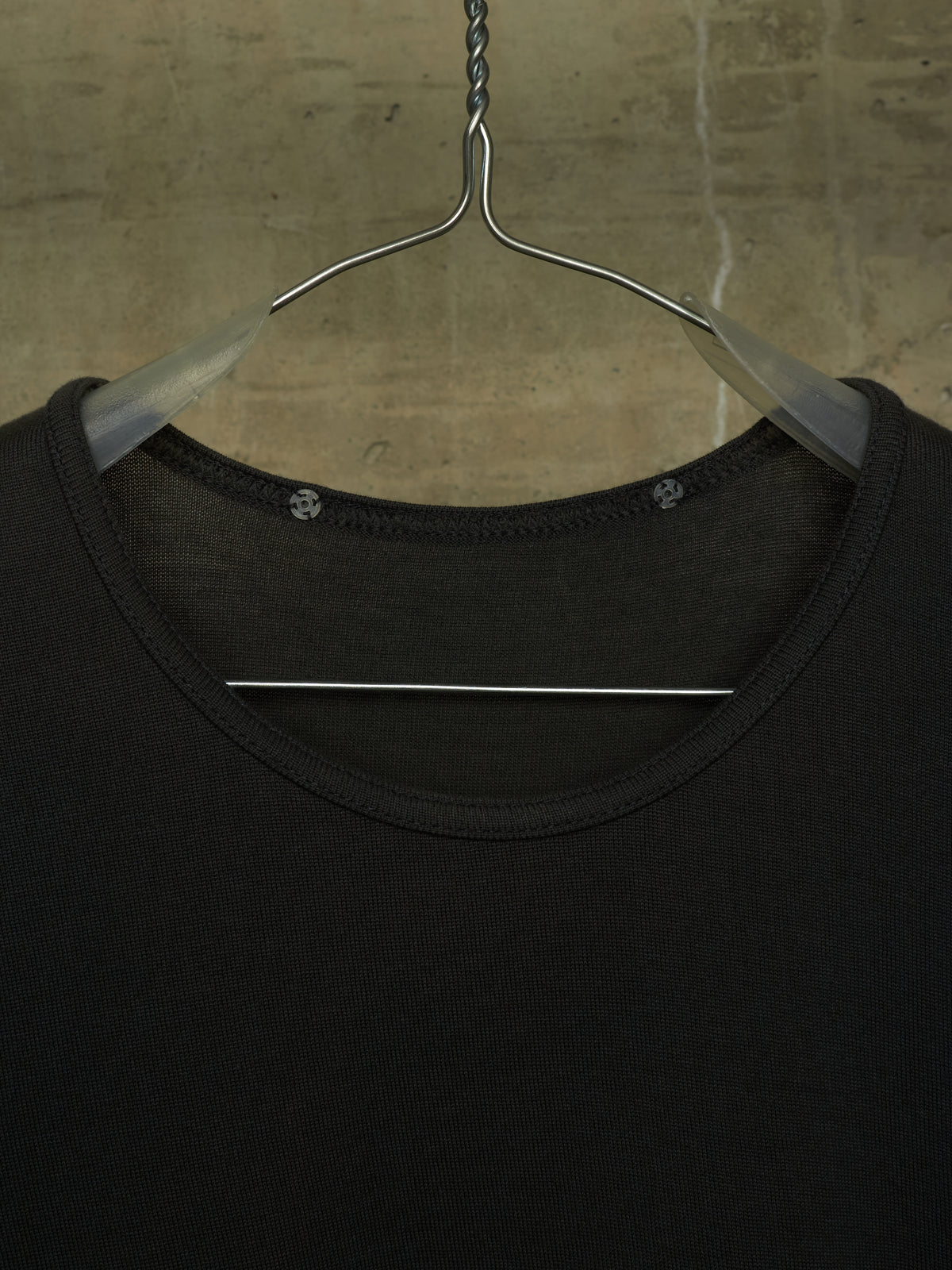 A close-up of a black crew-neck top hanging on a clear plastic hanger against a textured concrete wall.