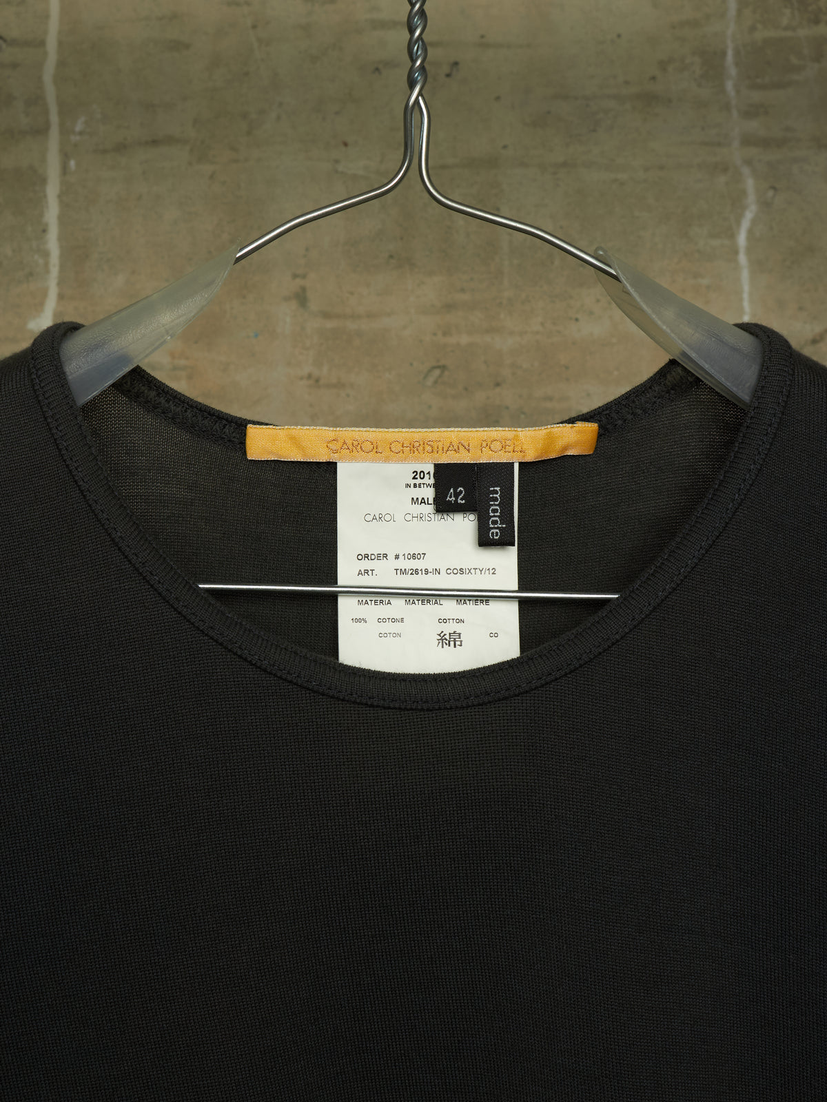 A black crewneck t-shirt hanging on a metal hanger against a textured concrete wall. The neck label shows the brand 'SACRED DESIGN CO.