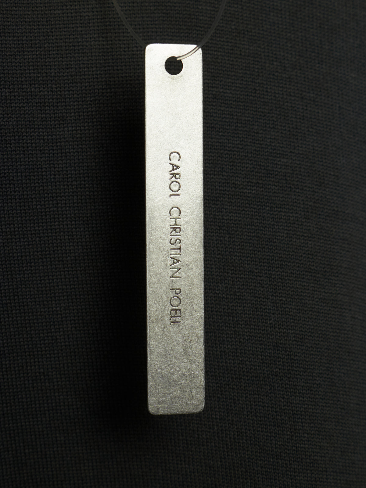 A rectangular silver metal tag engraved with the text 'CARIO CHRISTIAN POEL' hanging from a black cord against a black textured fabric background.