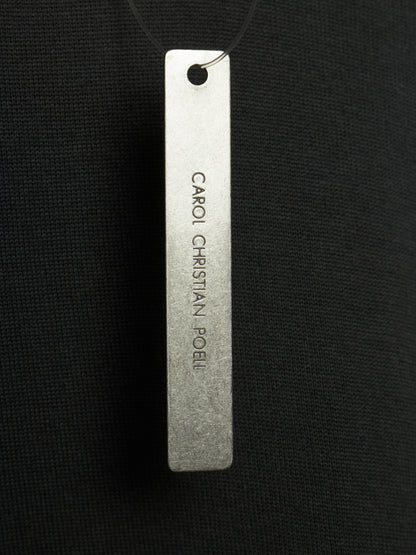 A rectangular silver metal tag engraved with the text 'CARIO CHRISTIAN POEL' hanging from a black cord against a black textured fabric background.