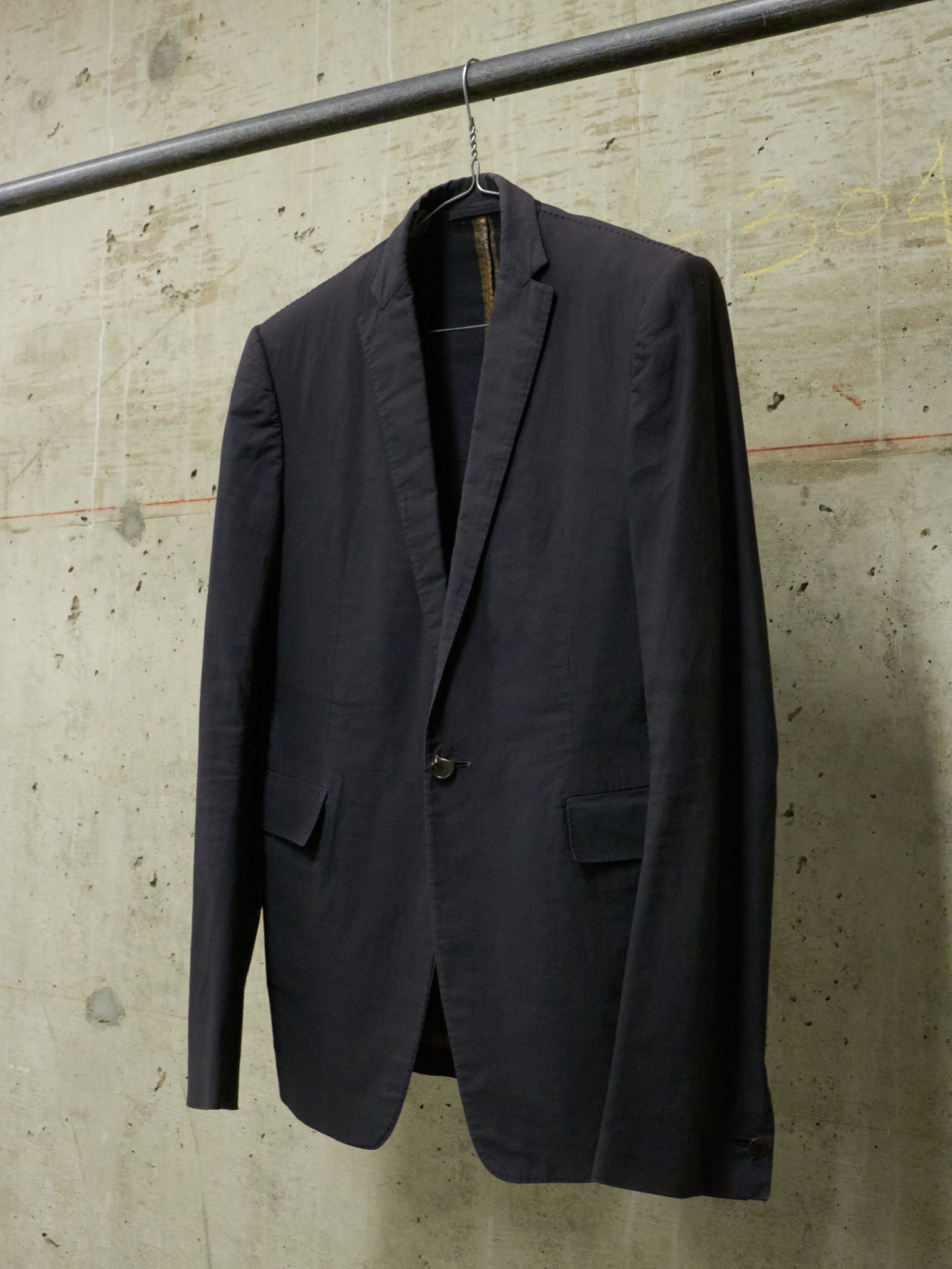 Dark gray blazer hanging on a hook against a textured concrete wall.