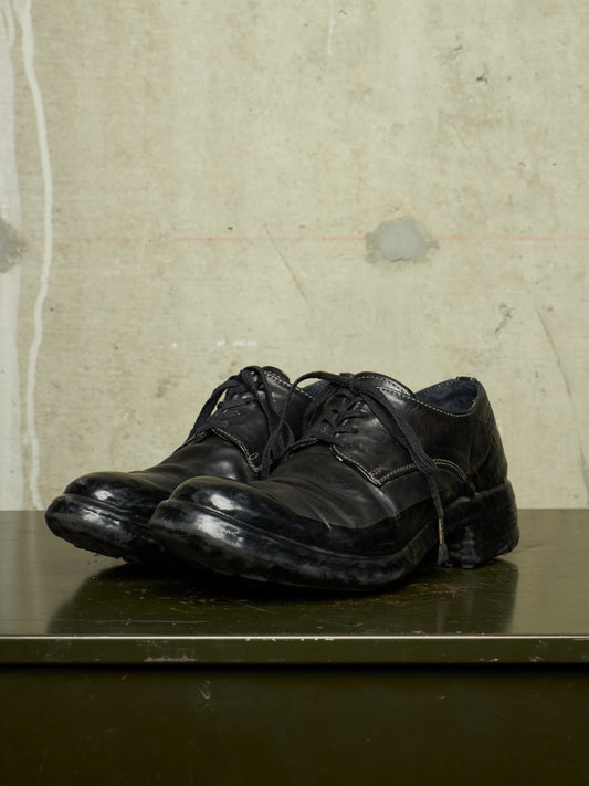 A pair of black leather lace-up dress shoes with a rugged, scuffed sole, displayed on a dark green surface against a textured concrete wall.