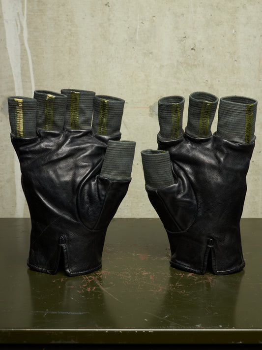 A pair of black leather fingerless gloves with ribbed gray cuffs and metallic gold accents, displayed on a dark green table against a textured beige wall.