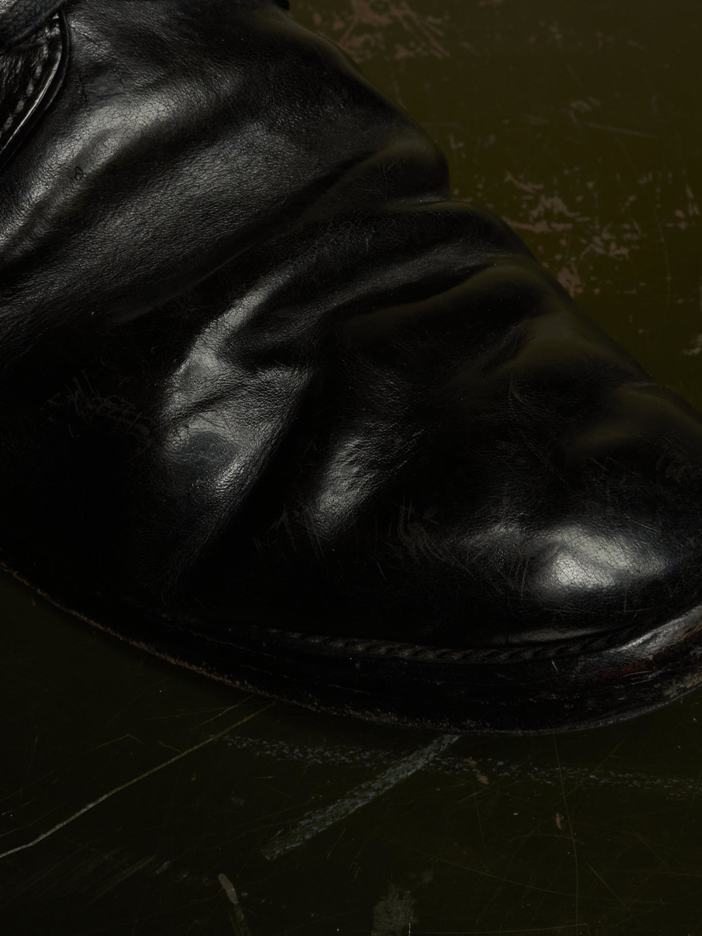 A close-up shot of a black leather boot, showing the textured, creased leather and a portion of the sole against a dark, distressed background.