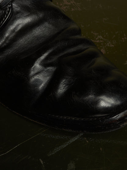 A close-up shot of a black leather boot, showing the textured, creased leather and a portion of the sole against a dark, distressed background.
