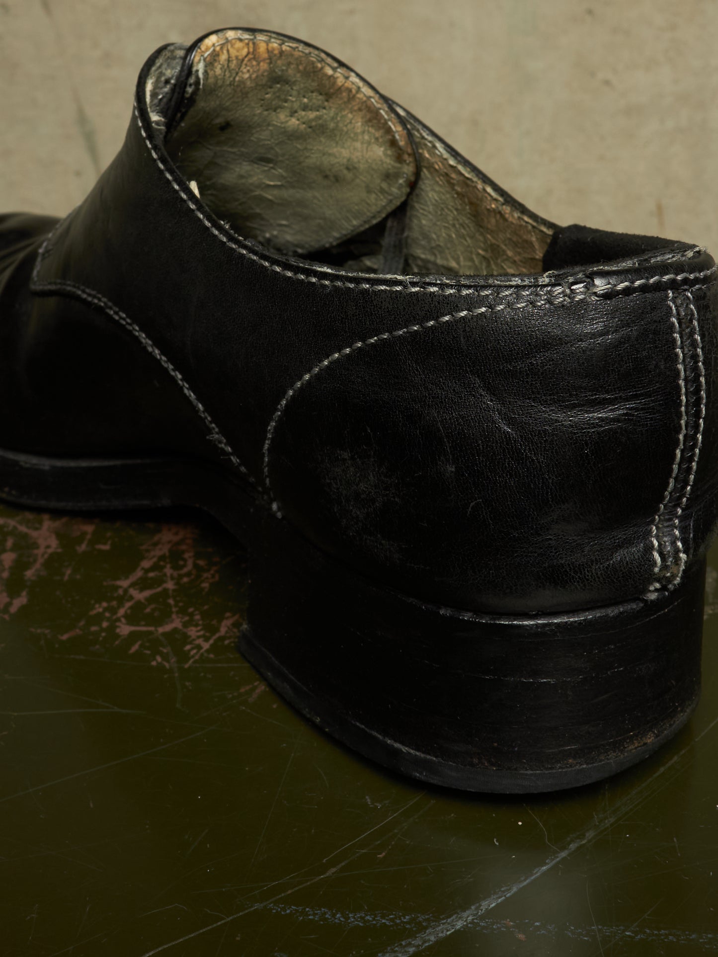 A close-up shot of a worn black leather shoe, showing the heel, sole, and interior lining, resting on a dark reflective surface.