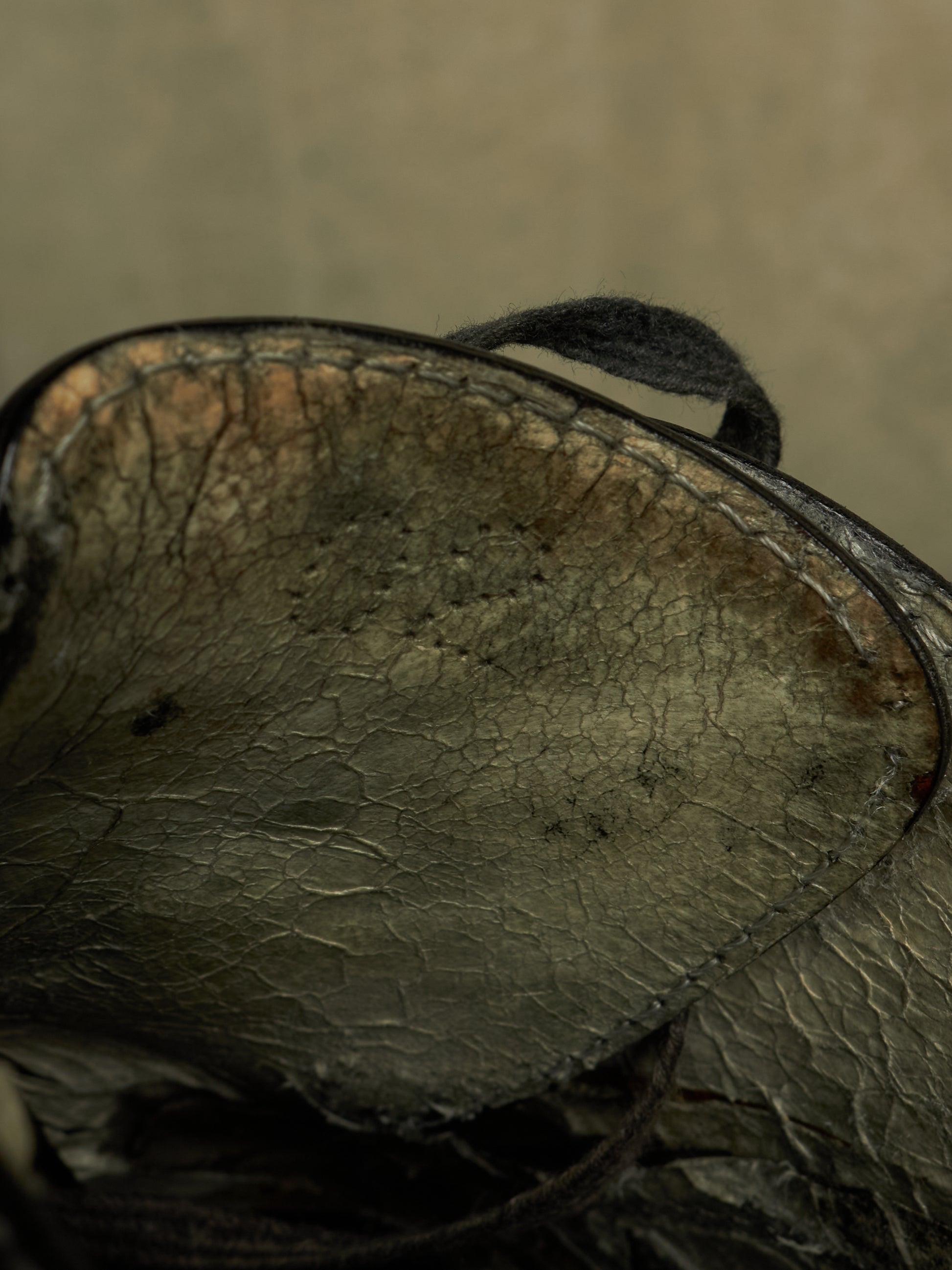 A close-up of a dark, distressed leather item showing the textured interior lining with visible cracks, scuffs, and a dark strap against a muted beige background.