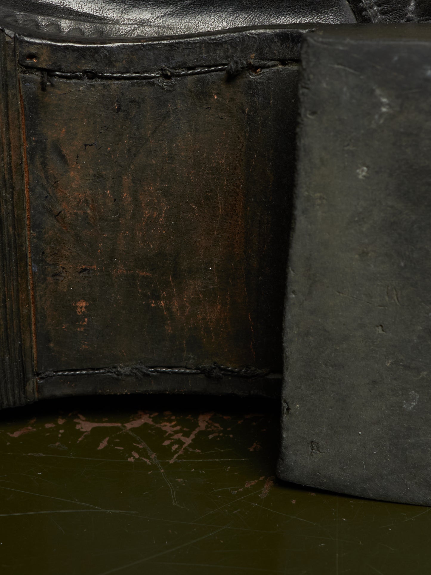 Close-up detail of a dark leather or leather-like product corner with heavy stitching, showing a worn brown interior panel and a dark gray side piece on a dark green surface.