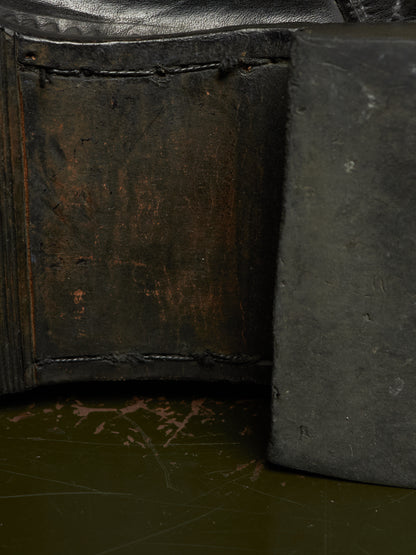 Close-up detail of a dark leather or leather-like product corner with heavy stitching, showing a worn brown interior panel and a dark gray side piece on a dark green surface.