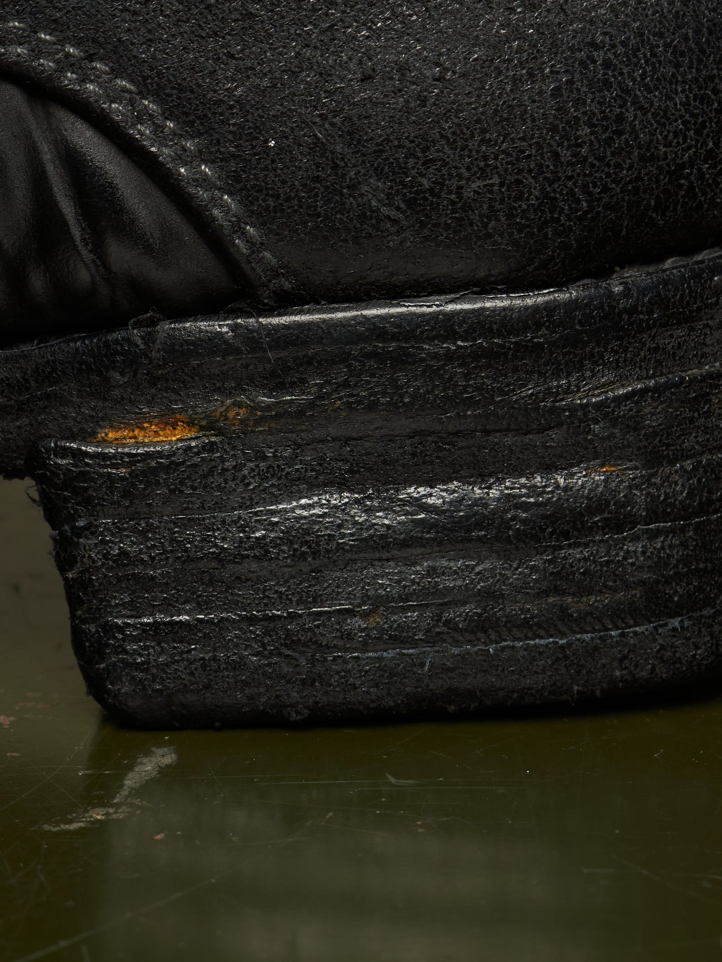Extreme close-up of a black leather shoe heel and sole, showing heavy scuffing, wear, and a small patch of exposed orange-brown rust on the stacked rubber sole.