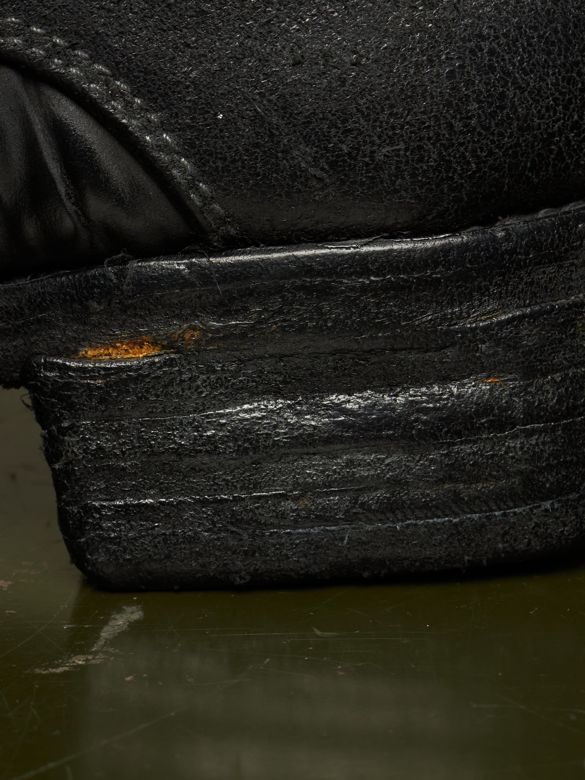Extreme close-up of a black leather shoe heel and sole, showing heavy scuffing, wear, and a small patch of exposed orange-brown rust on the stacked rubber sole.