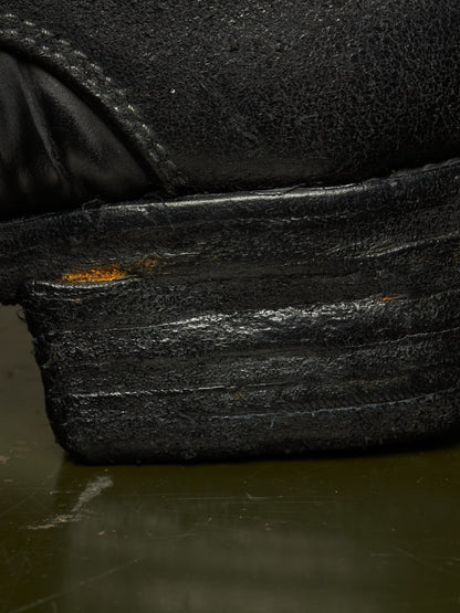 Extreme close-up of a black leather shoe heel and sole, showing heavy scuffing, wear, and a small patch of exposed orange-brown rust on the stacked rubber sole.