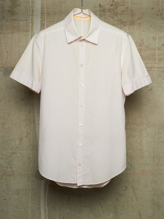 A short-sleeve button-up shirt in a very pale blush pink or off-white hue, hanging on a metal hanger against a textured concrete wall.