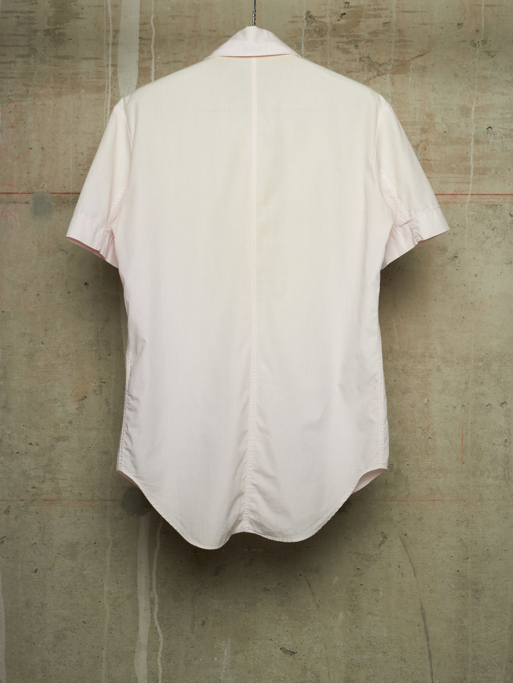 A light pink short-sleeve button-up shirt shown from the back, hanging against a textured concrete wall.