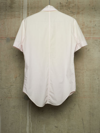 A light pink short-sleeve button-up shirt shown from the back, hanging against a textured concrete wall.