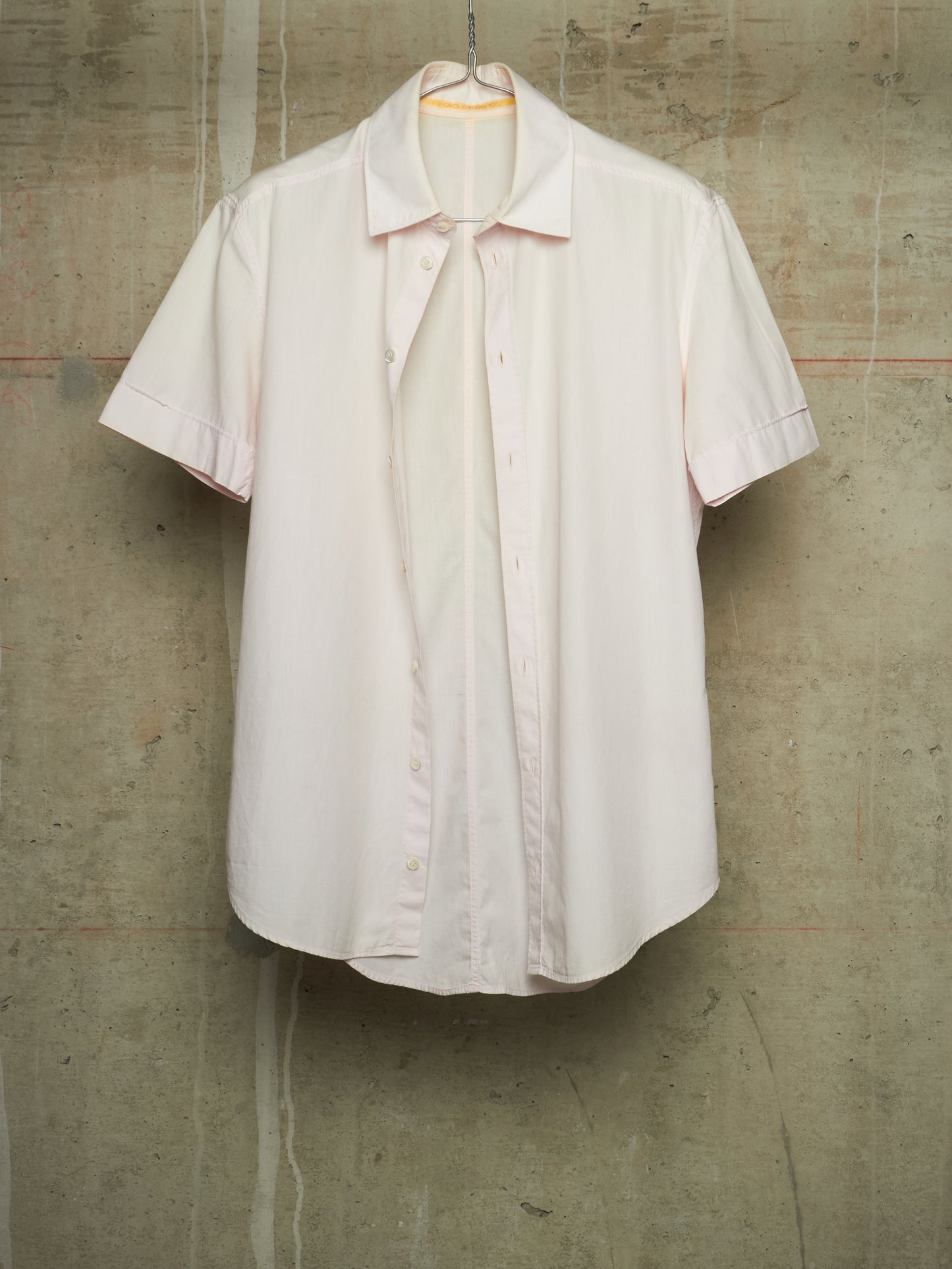 A short-sleeve button-up shirt in a very pale blush pink or off-white hue, hanging against a textured concrete wall.