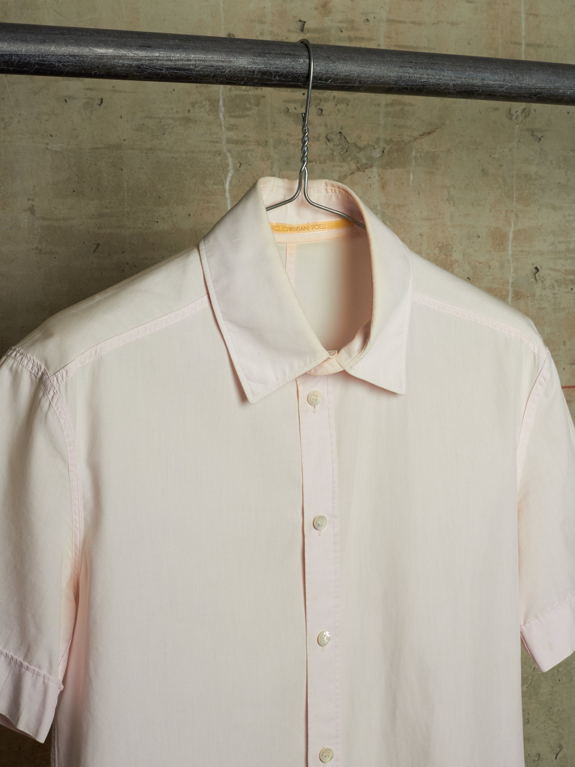 A pale blush pink short-sleeve button-up shirt hanging on a black metal hanger from a dark industrial rod, set against a textured concrete wall.