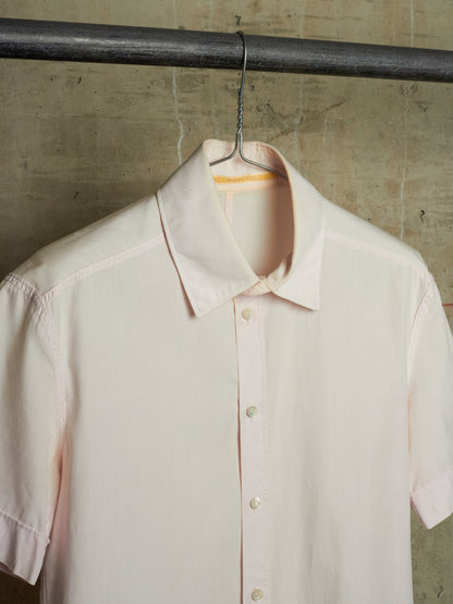 A pale blush pink short-sleeve button-up shirt hanging on a black metal hanger from a dark industrial rod, set against a textured concrete wall.