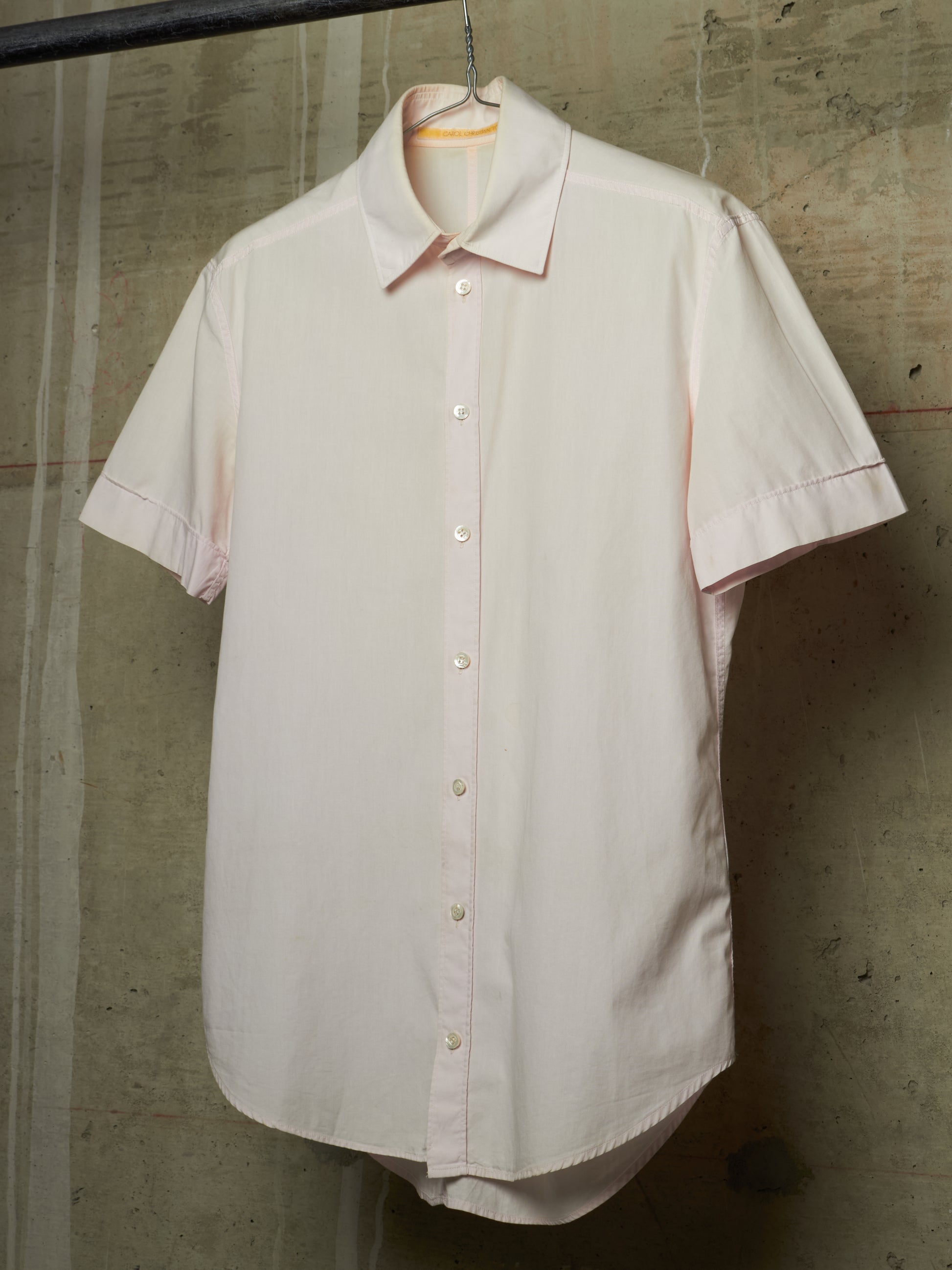 A light pink short-sleeve button-up shirt hanging on a black hanger against a textured concrete wall.