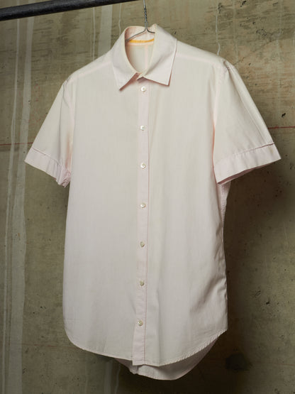 A light pink short-sleeve button-up shirt hanging on a black hanger against a textured concrete wall.